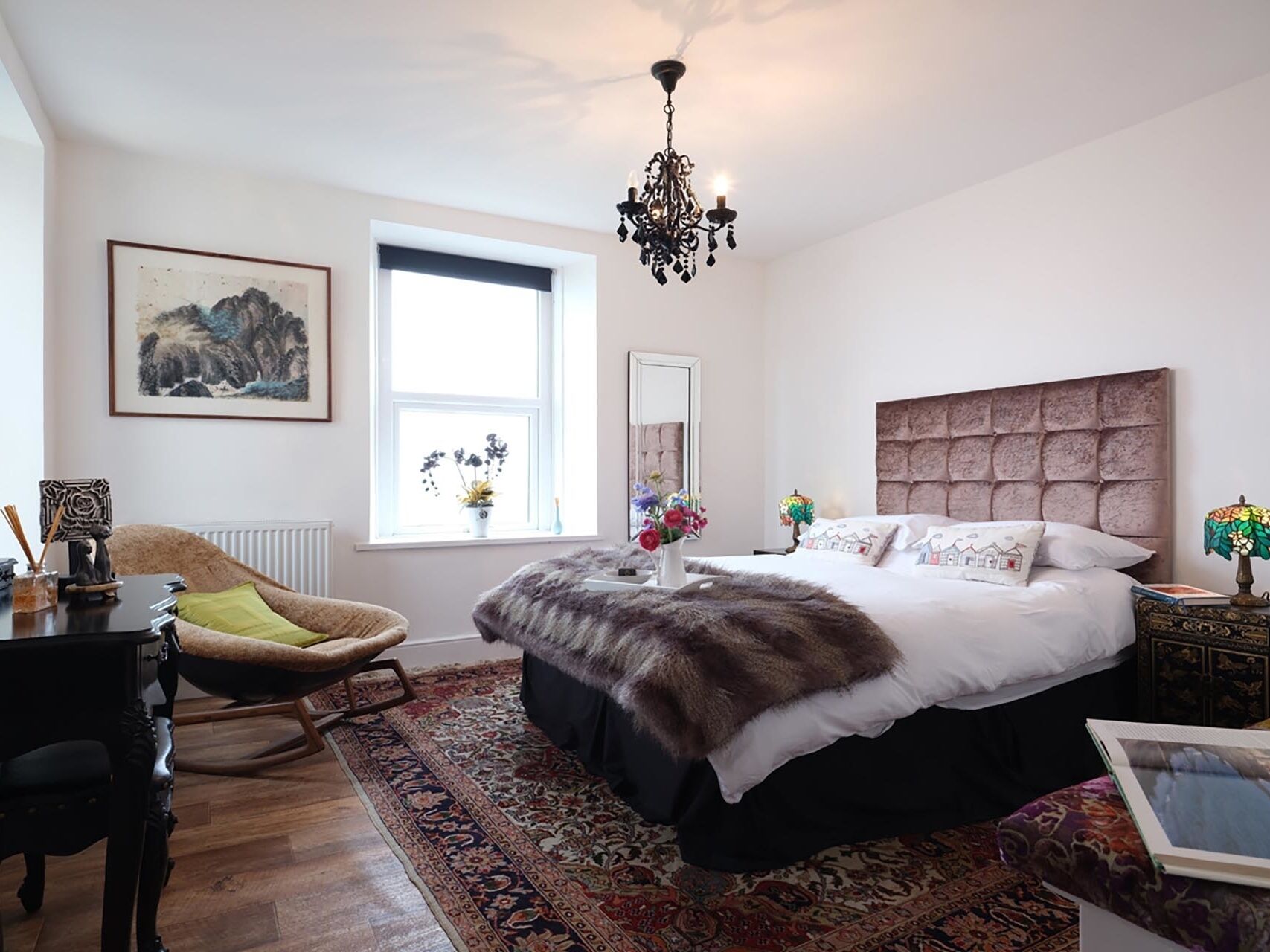 A bedroom with a large bed, a chandelier, a chair, a table, and a window with a view of the outdoors.