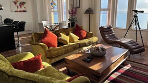 Un salón con un sofá amarillo, un sillón de cuero marrón, una mesa de centro de madera y vistas al mar a través de grandes ventanales.