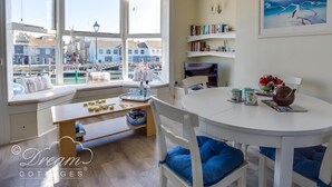 A cozy room with a bay window overlooking a harbor, a dining table with chairs, a bookshelf, and a painting on the wall.