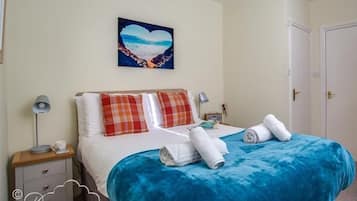 Ein ordentlich bezogenes Bett mit blauer Tagesdecke und Karokissen, eine Nachttischlampe und ein herzförmiges Bild an der Wand.