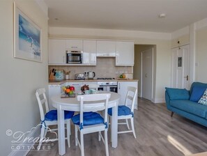 Una cocina compacta con armarios blancos, un microondas y una estufa a gas. Hay una mesa de comedor con cuatro sillas y un sofá azul en la sala.