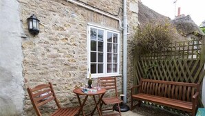 A stone cottage with a thatched roof, a wooden bench, and a small table with chairs. - Bramble Cottage (Bridport)