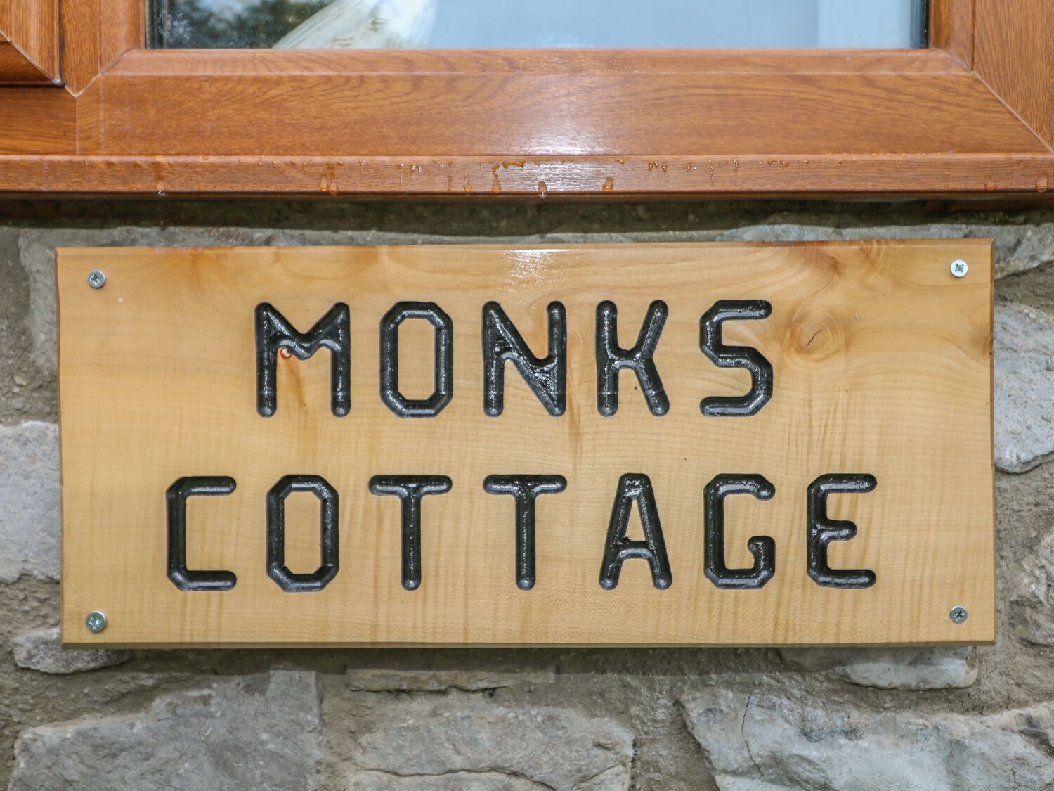 A wooden sign with the text "MONKS COTTAGE" mounted on a stone wall.