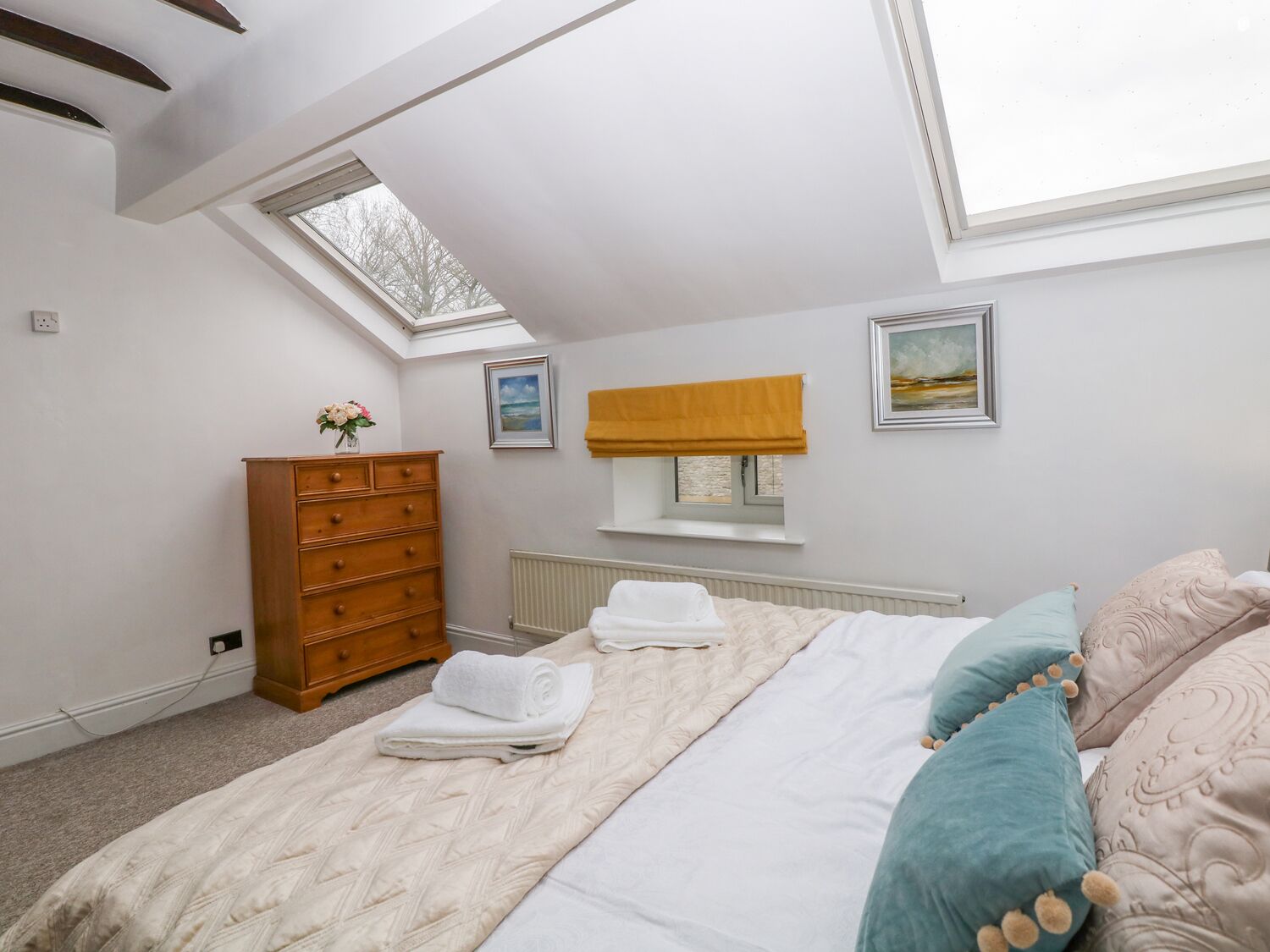 A bedroom with a skylight, a wooden dresser, a bed with pillows, and a framed picture on the wall.