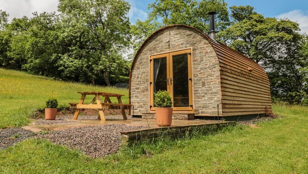 A small stone cabin with a wooden door and windows, a picnic table, and potted plants in a grassy area. - Dingle Den (Hereford)