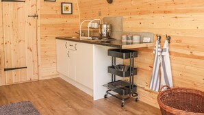 A compact kitchen with wooden walls, a sink, a stove, and a small countertop. There are two framed pictures on the wall, a basket, and a towel rack. - Dingle Den (Hereford)