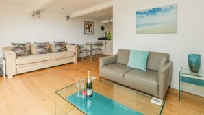 A modern living room with a glass coffee table, two sofas, a dining area with a small table and chairs, and a kitchen in the background. - Burton Bay (Bridport)