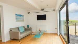 A modern living room with a glass coffee table, a sofa with a cushion, a mounted TV, and a view of the outdoors. - Burton Bay (Bridport)