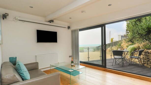 A modern living room with a glass coffee table, a grey sofa with teal cushions, a flat-screen TV mounted on the wall, and a large sliding glass door leading to an outdoor patio with a wooden deck and a view of the beach. - Burton Bay (Bridport)