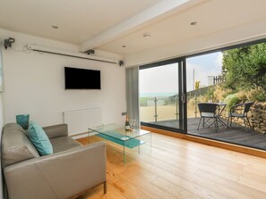 Ein modernes Wohnzimmer mit Glastisch, grauem Sofa mit türkisfarbenen Kissen, einem flachbildfernseher an der Wand und einer großen Schiebetür zu einer Terrasse mit Holzdeck und Blick auf den Strand.