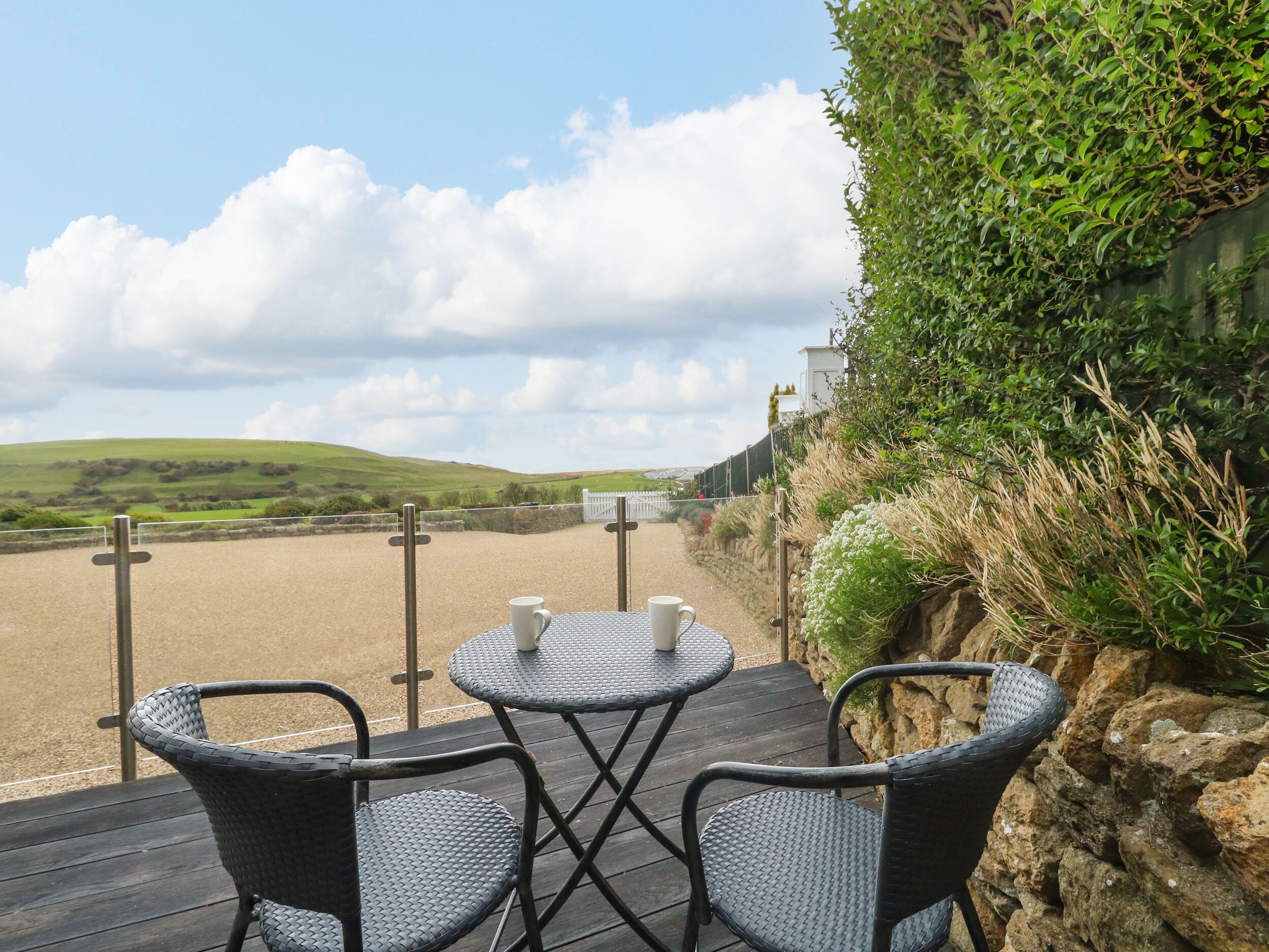 A terrace with wicker furniture and a table, overlooking a scenic landscape with hills and a fence.