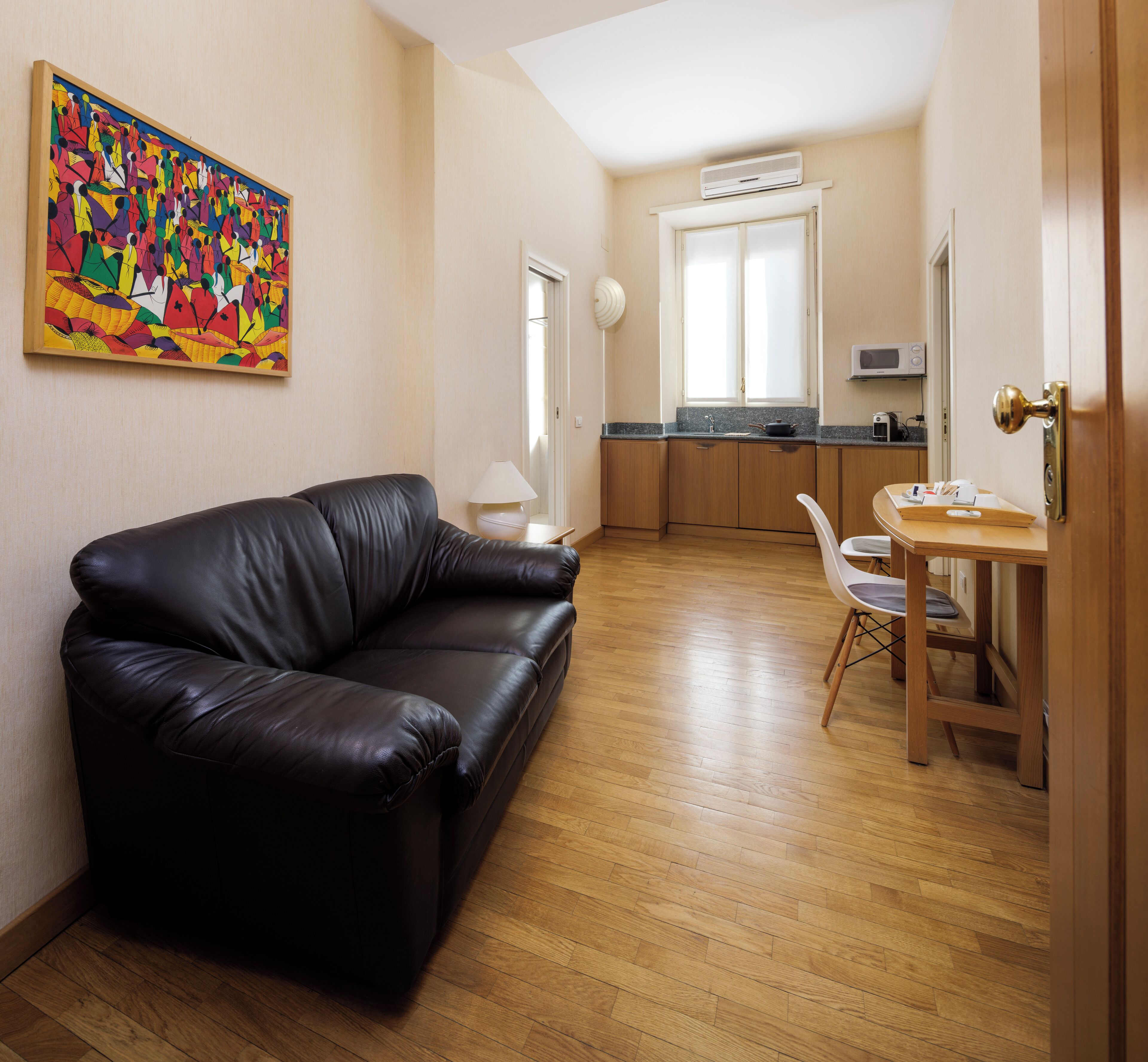A modern living room with a black leather sofa, a dining area with a wooden table and chairs, a kitchen with wooden cabinets and a microwave, and a colorful abstract painting on the wall.