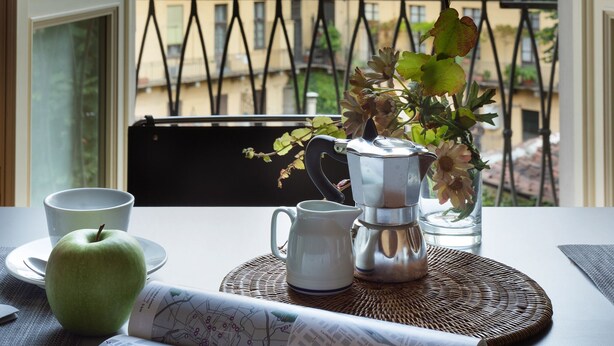 Ein Tisch mit einem grünen Apfel, einer weißen Tasse, einer silbernen Kaffeekanne und einer Vase mit Blumen.