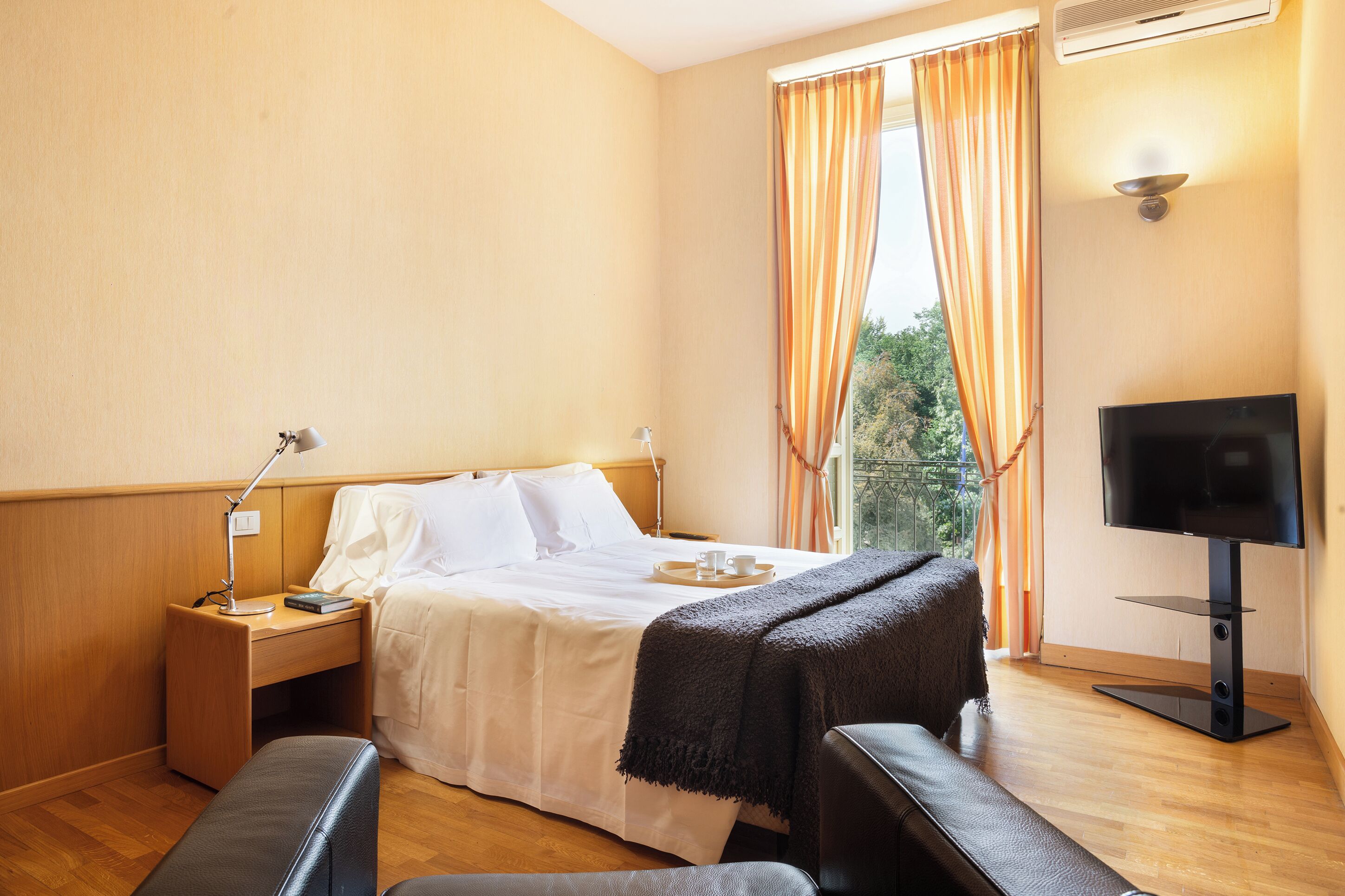 A hotel room with a bed, bedside table, lamp, TV, and a view of greenery through the window.