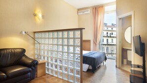 Ein Zimmer mit Holzboden, einem schwarzen Ledersofa, einem Bett mit weißer Bettdecke, einem Fernsehschrank und einem Fenster mit Vorhängen.