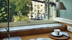 Ein Schreibtisch mit einer Lampe, einer Tasse und einem offenen Buch neben einem Fenster mit Blick auf Gebäude.