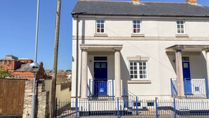 A two-story building with a blue door and windows, a stone wall, and a metal fence. - Ashwood (Weymouth)