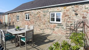 A stone building with a tiled roof, a patio with a table and chairs, and a small window.