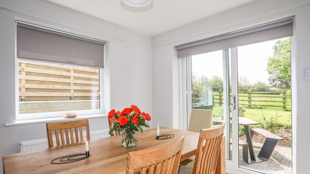 A dining area with a wooden table, chairs, a vase with flowers, and a view of the garden through large windows.