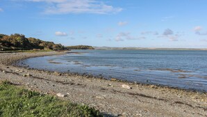 A coastal landscape with a pebbly beach, calm water, and distant hills.