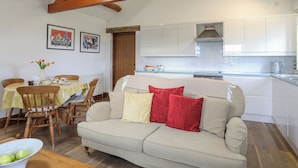 A cozy living room with a sofa, wooden chairs, a dining table with a vase of flowers, and a kitchen in the background.