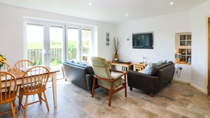 A living room with a dining area, a sofa, a TV, and a window with a view. - Farm Cottage (Cemaes Bay)