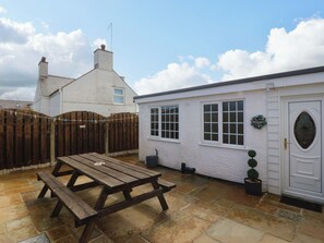 A patio with a wooden table and benches, a white building with a door and windows, and a wooden fence. - Farm Cottage (Cemaes Bay)