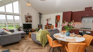 A modern living room with a fireplace, grey sofas, a dining table with orange chairs, and a kitchen with red cabinets. - Meadowside (Penzance)