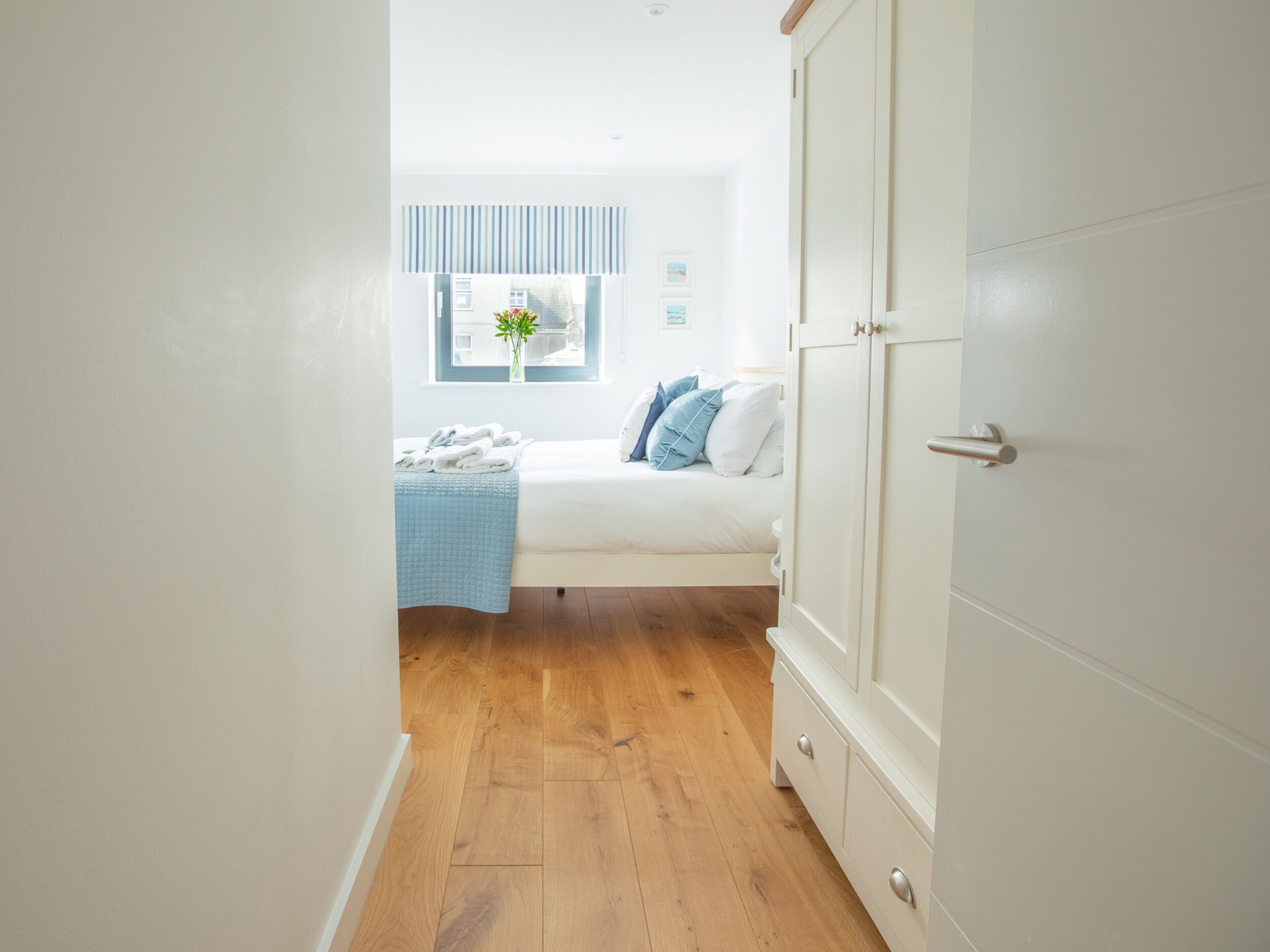 A bedroom with a bed, a window with blinds, a bedside table with a vase of flowers, and a wardrobe.