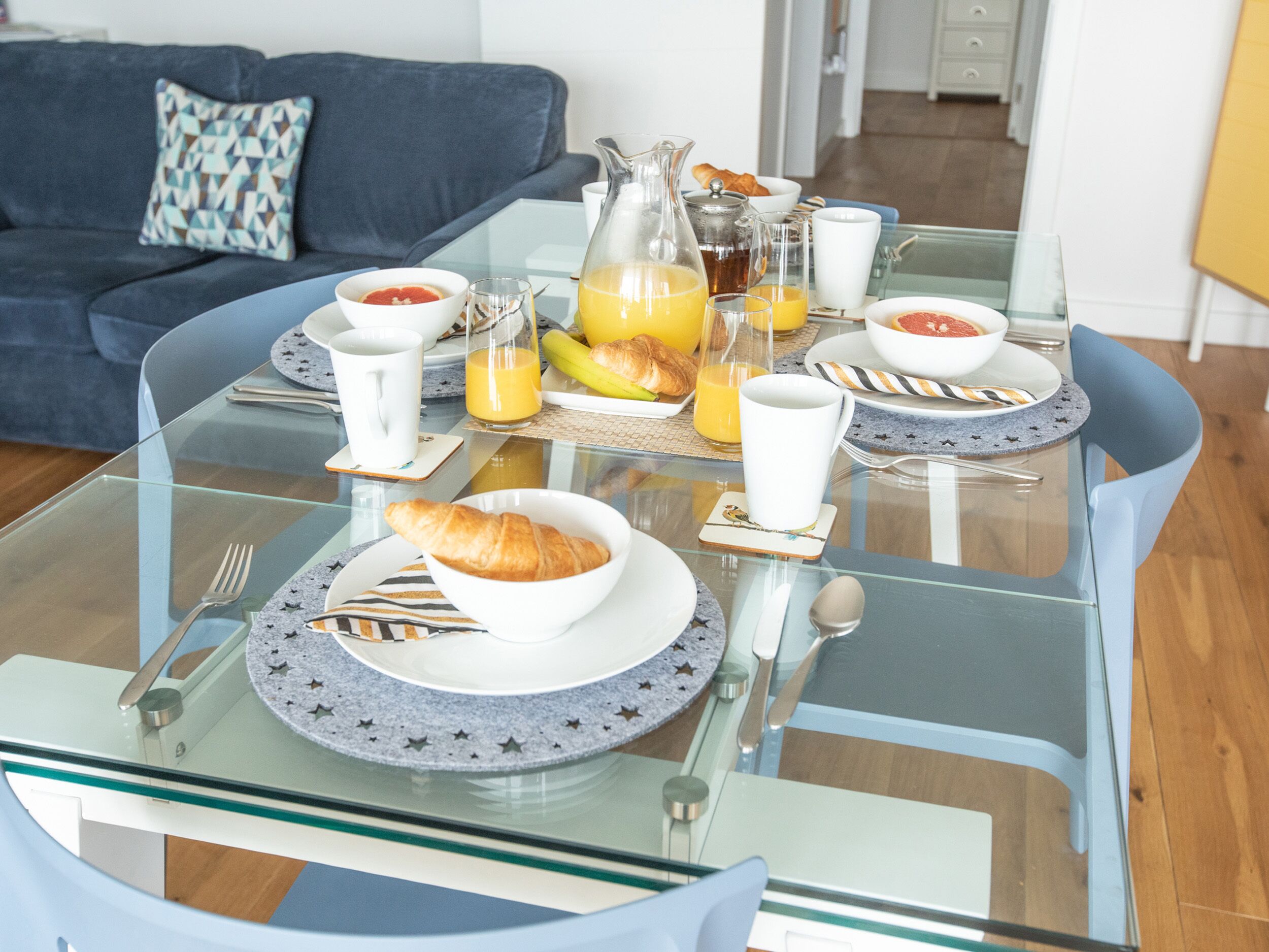 Une table de petit-déjeuner dressée avec des assiettes, des bols, des croissants et des verres de jus d’orange.