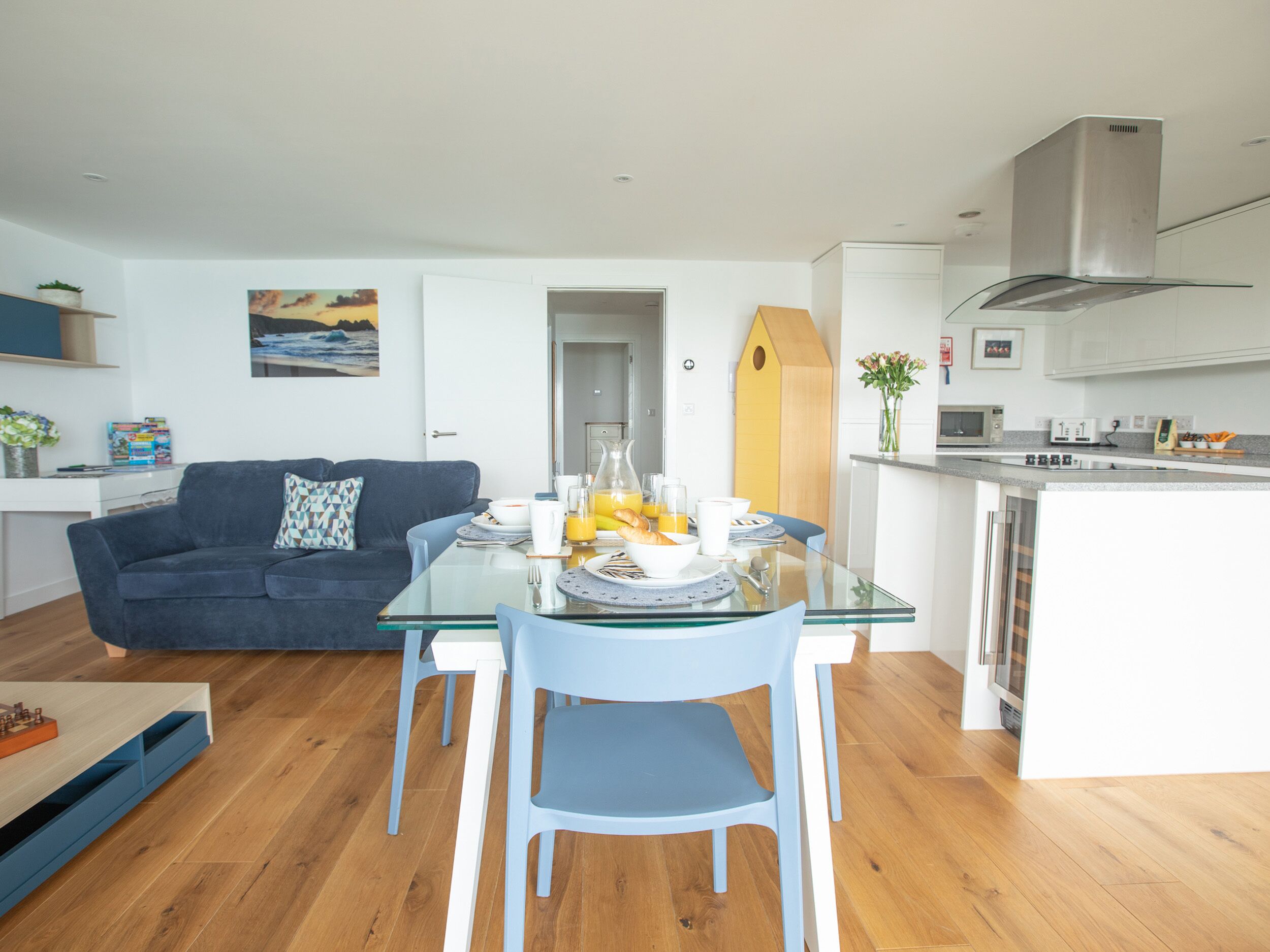 Un espace de vie moderne comprenant une table à manger en verre, des chaises bleues, un canapé bleu foncé et un coin cuisine.