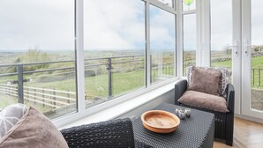 A conservatory with large windows offering a view of the countryside. Inside, there is a wicker sofa with cushions and a wooden bowl on a woven table. - Frondeg (Bangor)