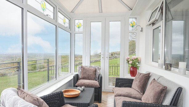 A sunroom with wicker furniture, a view of the countryside, and a red vase with flowers. - Frondeg (Bangor)