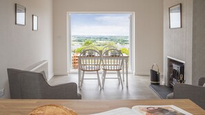 A modern living room with a dining area, a fireplace, and a view of a green landscape through a large window. - Frondeg (Bangor)