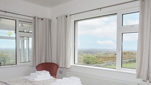 A bedroom with a bed, a chair, and a view of a landscape through large windows. - Frondeg (Bangor)
