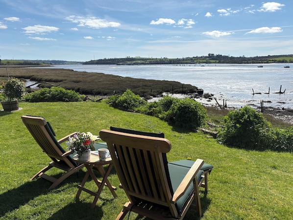 A grassy area with two wooden chairs and a table, overlooking a body of water with boats.