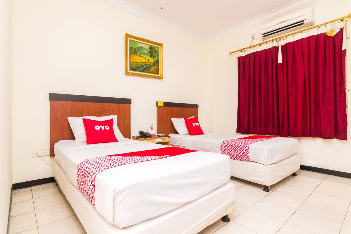 Une chambre d’hôtel avec deux lits, chacun agrémenté de draps blancs et d’oreillers rouges, un tableau au mur et un rideau rouge.