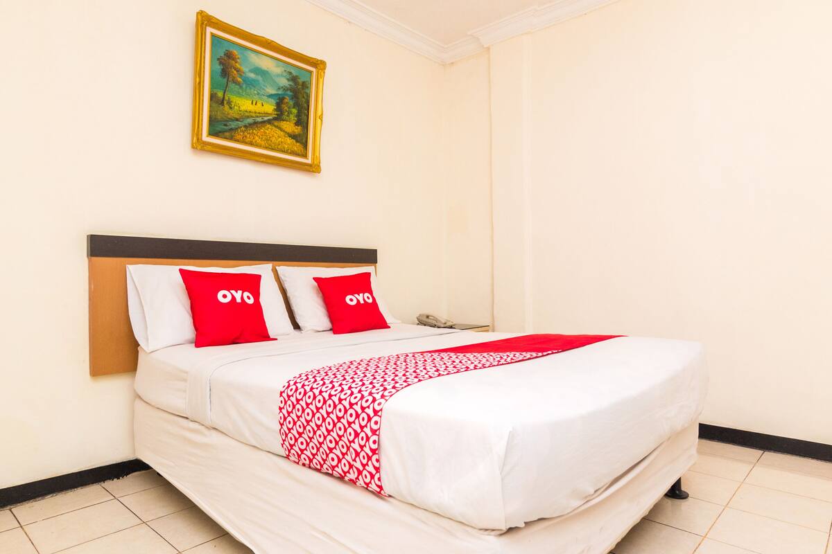 Un lit bien fait, avec une literie blanche et rouge, deux oreillers rouges sur lesquels est inscrit le mot « OYO », et un tableau accroché au mur au-dessus du lit.