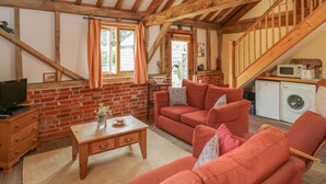 A cozy living room with a brick wall, wooden beams, a sofa set, a television, a table with a vase, and a washing machine.