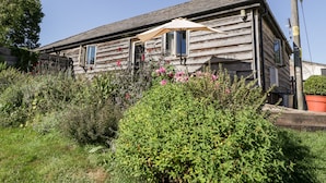 A rustic wooden house with a sloped roof, a porch with a beige umbrella, and a garden with various plants and flowers.