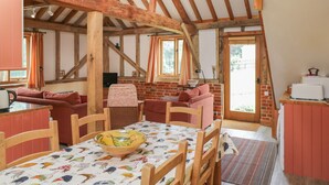 A rustic room with exposed wooden beams, a brick fireplace, a dining table with chairs, and a kitchen area with a refrigerator and a microwave.