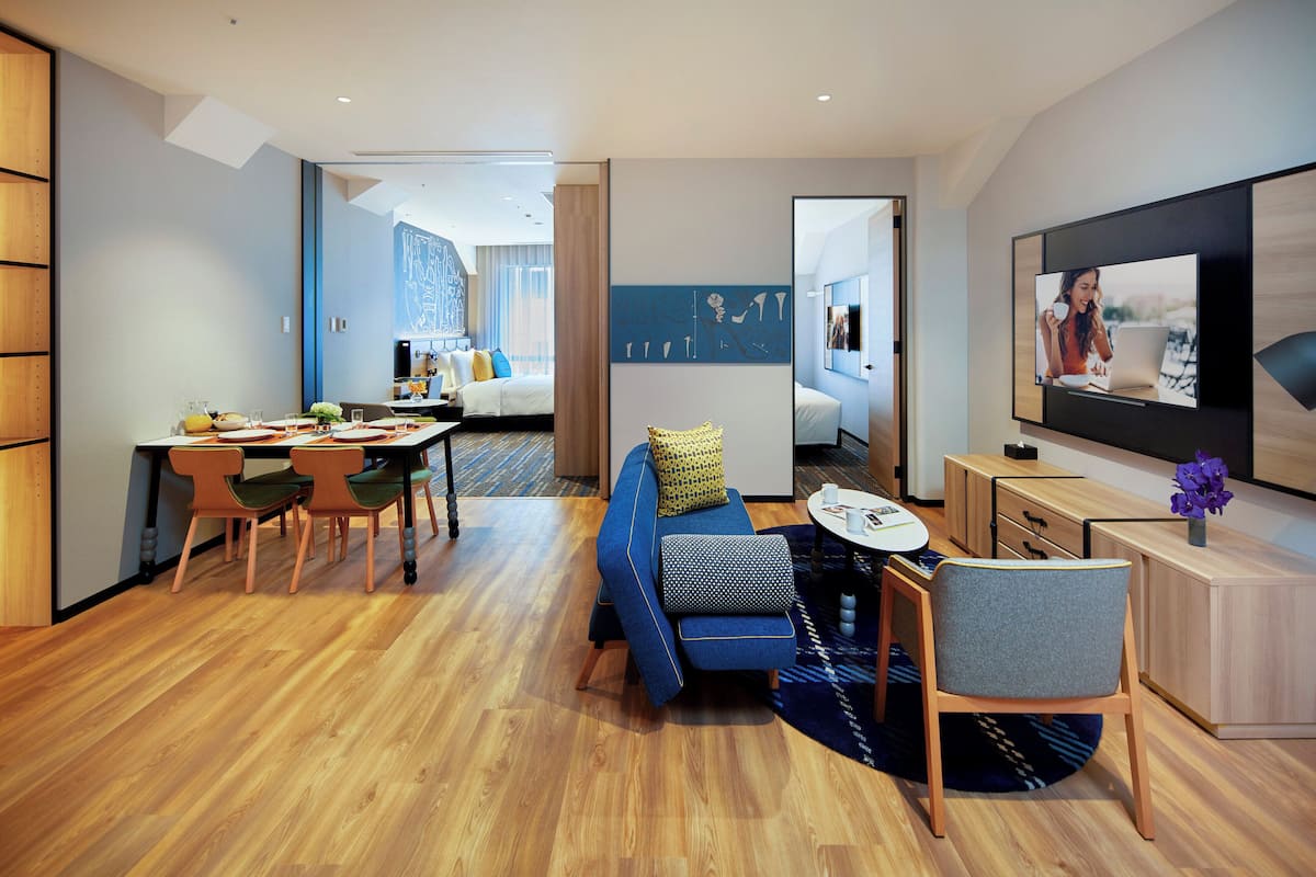 A modern hotel room with a dining area, a sofa, a TV, and a wooden floor.