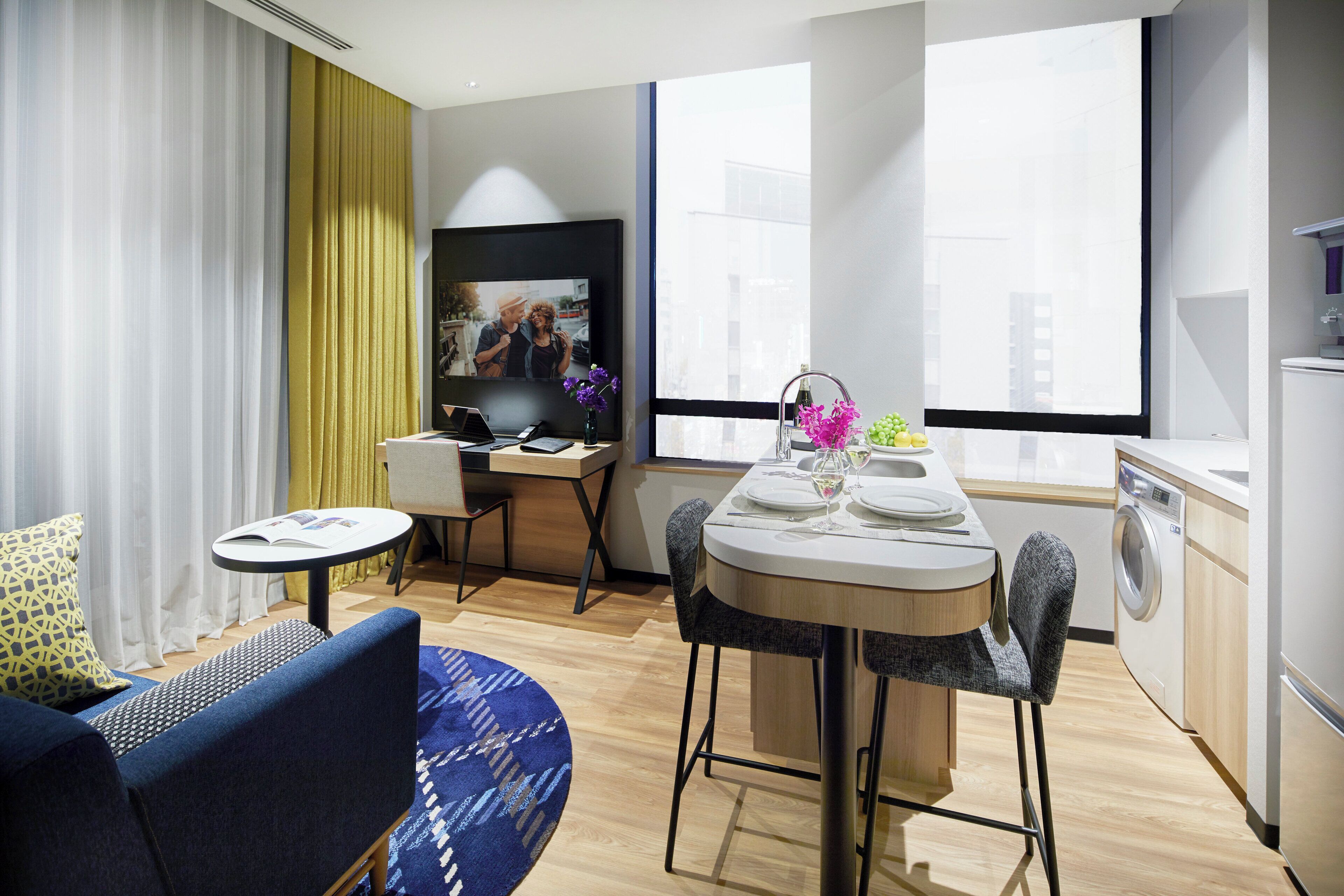 A modern hotel room with a dining area, a sofa, a small table, and a washing machine.