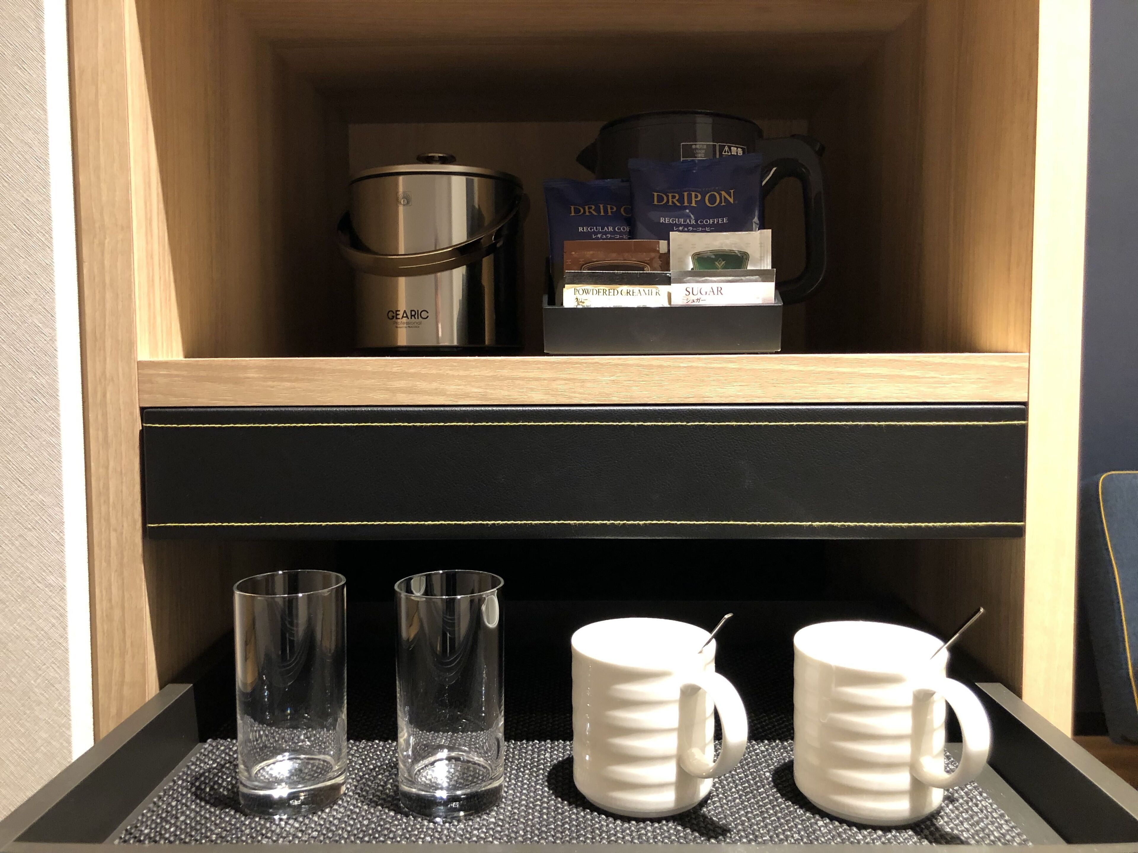 A minibar with a GEARIC coffee maker, DRIPON coffee bags, a powdered creamer box, and sugar. There are three clear glasses and two white mugs with handles.