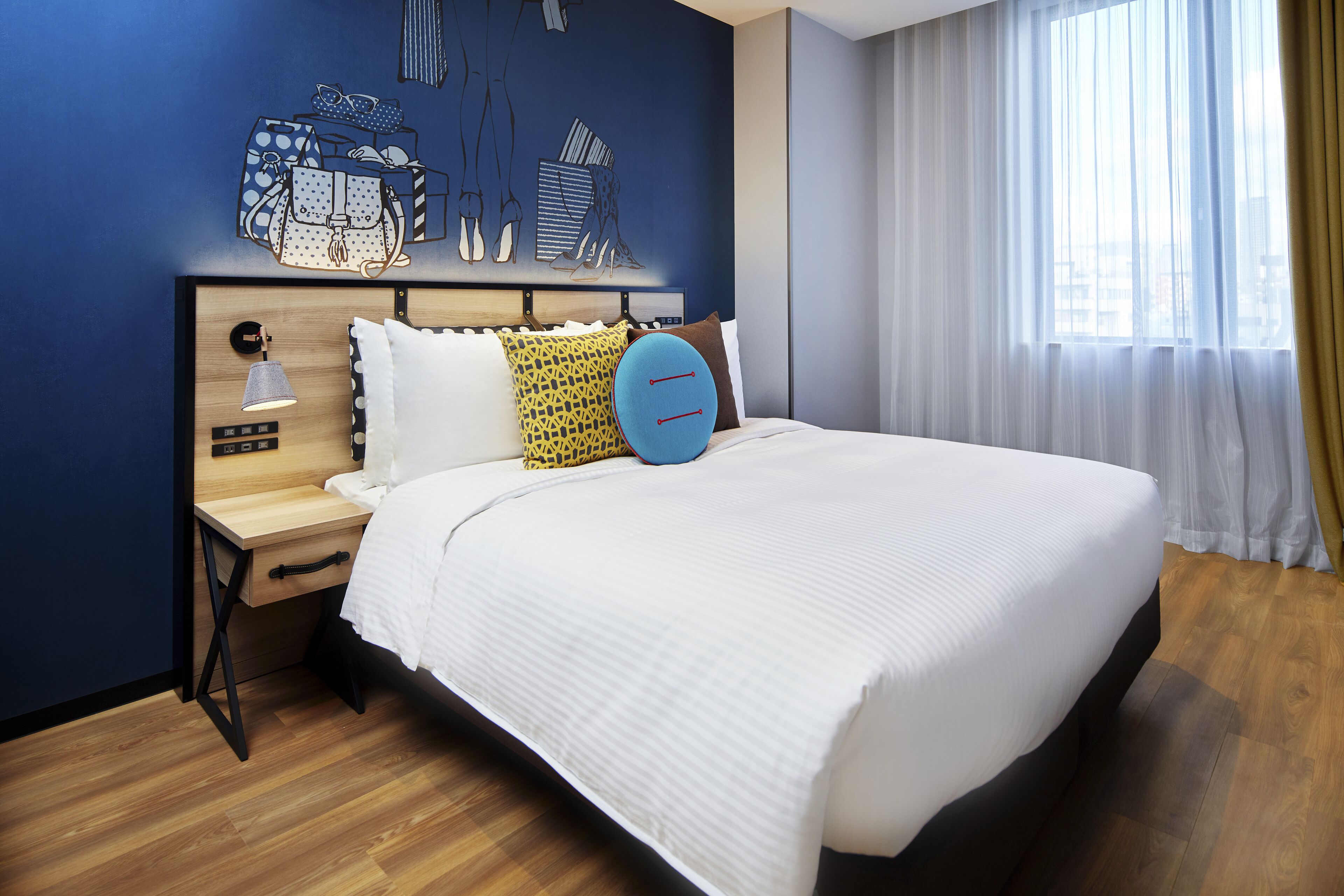 A modern hotel room with a large bed, a wooden nightstand, a desk, and a wall art piece depicting handbags and shoes.