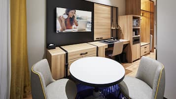 A compact room with a desk, two chairs, a round table, and a television mounted on the wall.