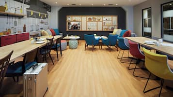 A modern dining area with wooden tables and colorful chairs, a kitchen with a chalkboard wall, and a large window providing natural light.