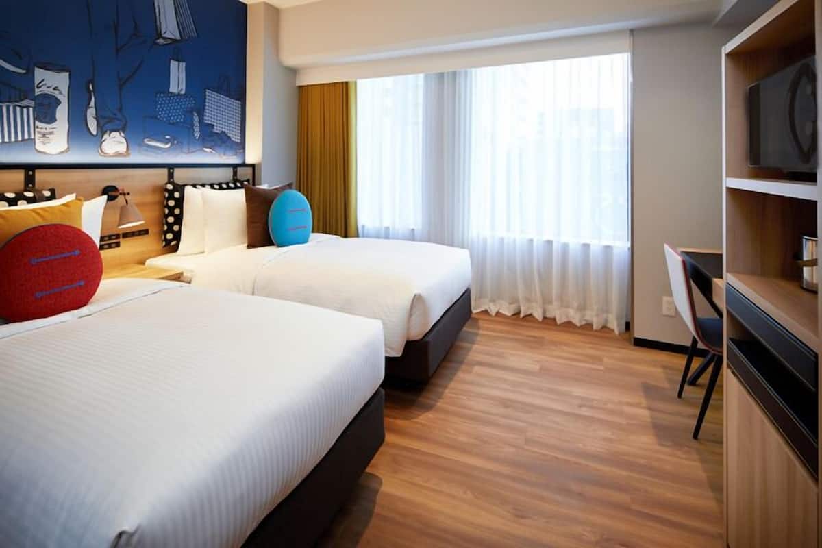 A hotel room with two beds, a desk, a television, and a large window with curtains.