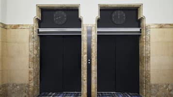 A pair of elevator doors with clocks above them, set in a stone wall with a patterned carpet.
