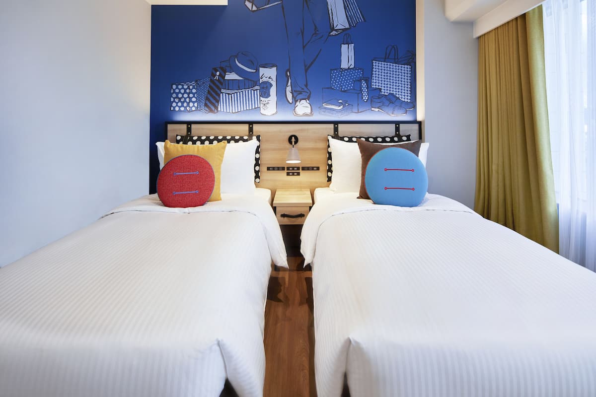 A hotel room with two single beds, a wooden headboard, and a large artwork depicting shopping bags and shoes on the wall.